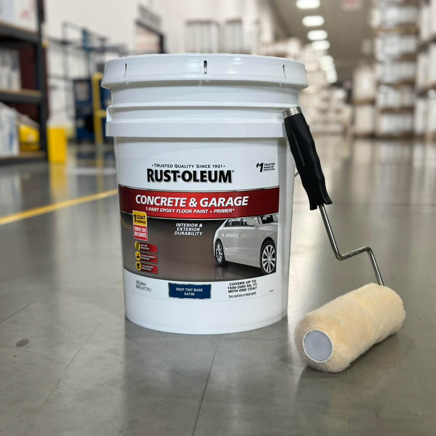 A labeled Rust-Oleum Concrete & Garage floor paint pail with a paint roller on a warehouse floor