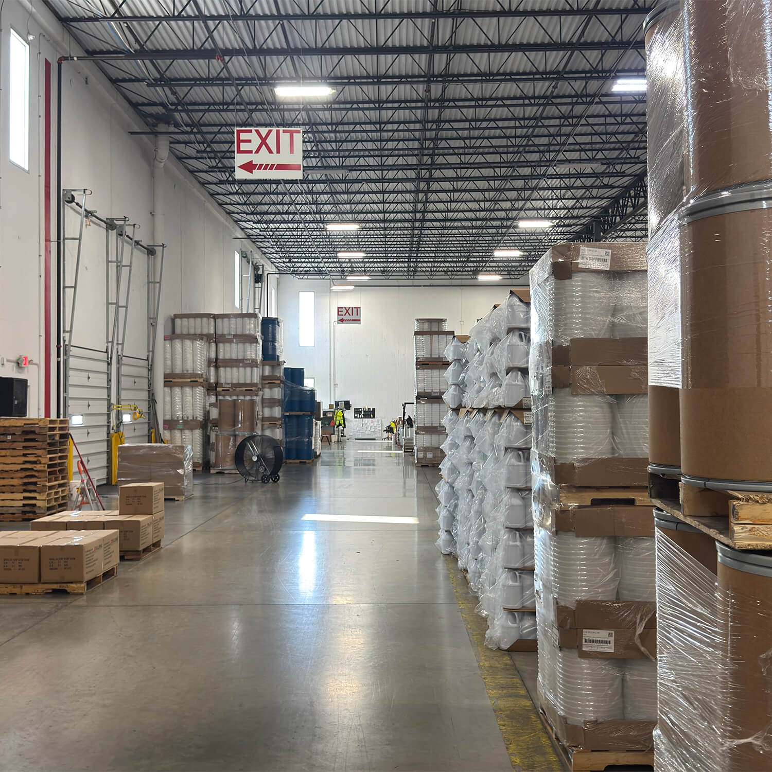 A long ILLING warehouse aisle with loading docks, staged pallets of pails and jugs, and fiber drums ready to ship